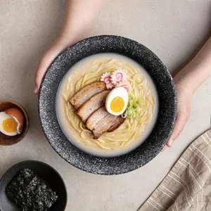 Main féminine présentant un bol de ramen avec un bouillon épais, des tranches de porc grillé, un œuf à la coque coupé en deux, et des tranches de narutomaki, avec des bols contenant un œuf et des algues séchées à côté