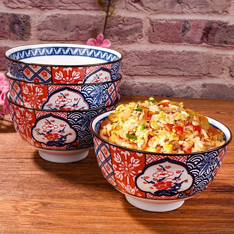 Trois bols en céramique empilés, présentant des motifs floraux et géométriques en rouge et bleu, avec une mise au point sur le bol supérieur. Ils sont placés sur une planche en bois avec des fleurs roses floues en arrière-plan