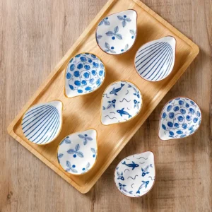 Des plats en céramique en forme de larme avec des motifs en bleu, présentés sur une table en bois. Les motifs incluent des feuilles, des lignes abstraites, et des poissons