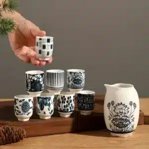 Une main tenant une petite tasse de saké en céramique décorée de motifs bleus et blancs, avec d'autres tasses et une cruche sur un plateau en bois en arrière-plan.