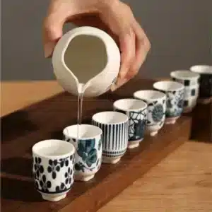 Une main verse du saké d'une cruche en céramique blanche dans une série de tasses assorties alignées sur un plateau en bois