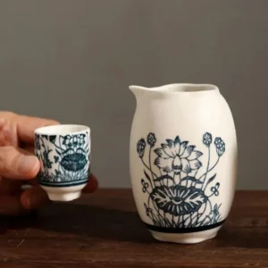 Une petite tasse de saké en céramique avec un motif floral bleu et blanc tenu à côté d'une cruche assortie sur un fond neutre en bois.